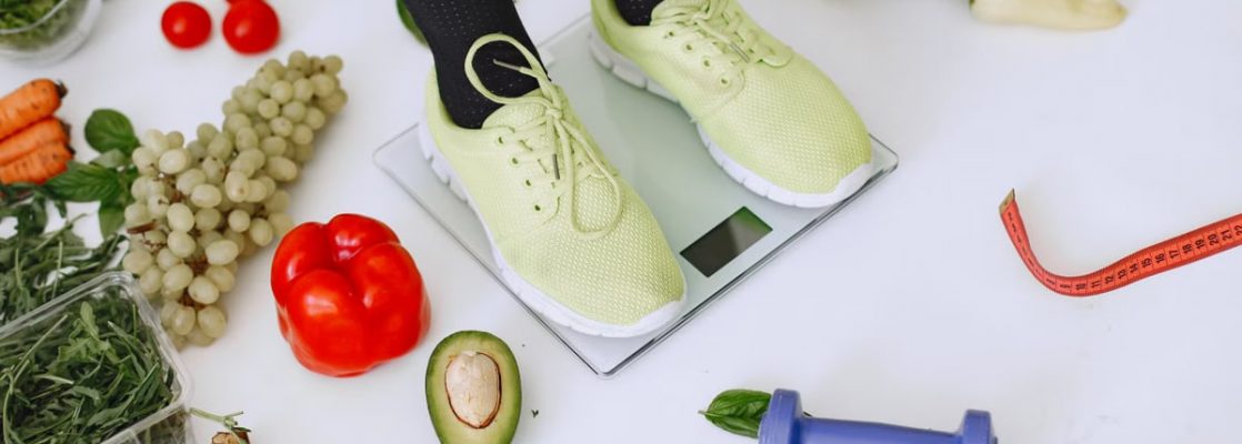 Person standing on scales surrounded by fresh produce and fitness equipment symbolizing a balanced lifestyle.