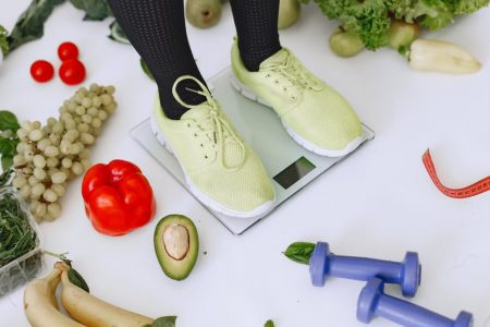 Person standing on scales surrounded by fresh produce and fitness equipment symbolizing a balanced lifestyle.
