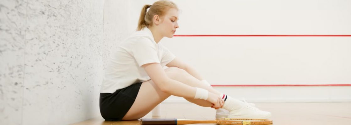 Side view of a young woman resting on a squash court, wearing sportswear and sitting with a racket.