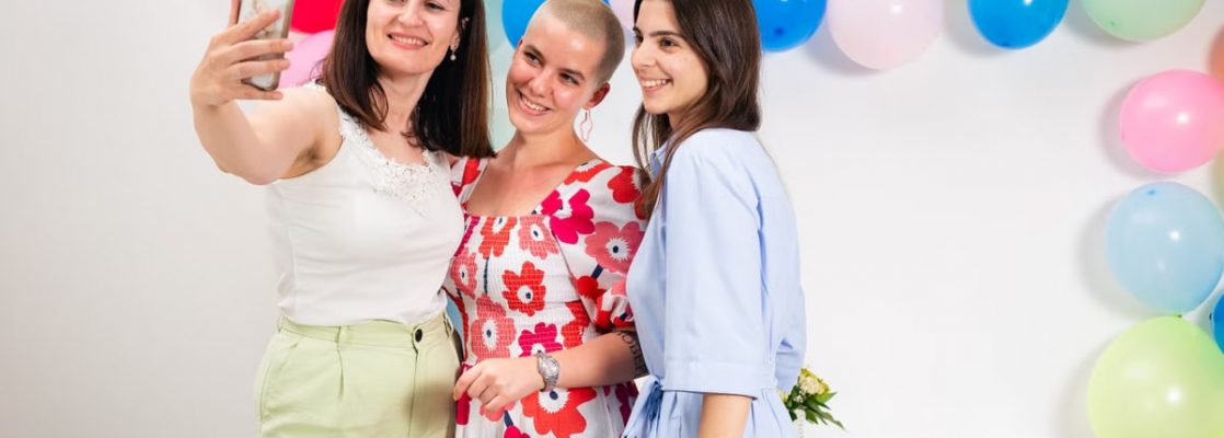 Three women joyfully taking a selfie against a colorful balloon backdrop indoors.