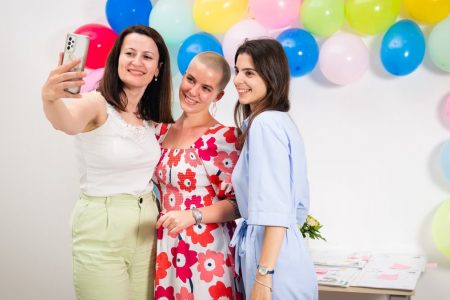 Three women joyfully taking a selfie against a colorful balloon backdrop indoors.